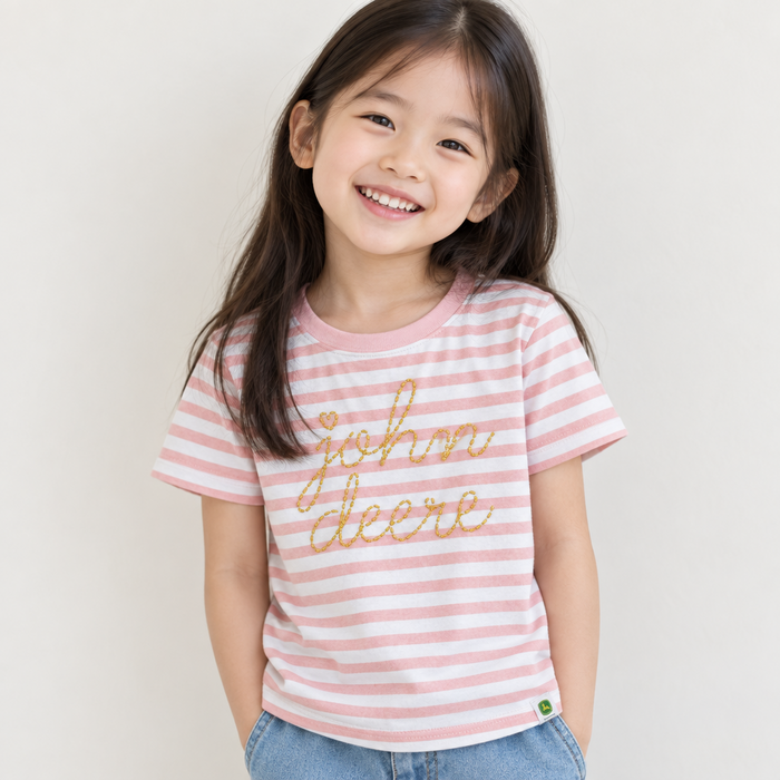 A young girl smiling while wearing a pink and white striped t-shirt with 'john deere' written in gold glitter script on the front, paired with light blue jeans, against a plain light background. John Deere Girls Stripe Tee