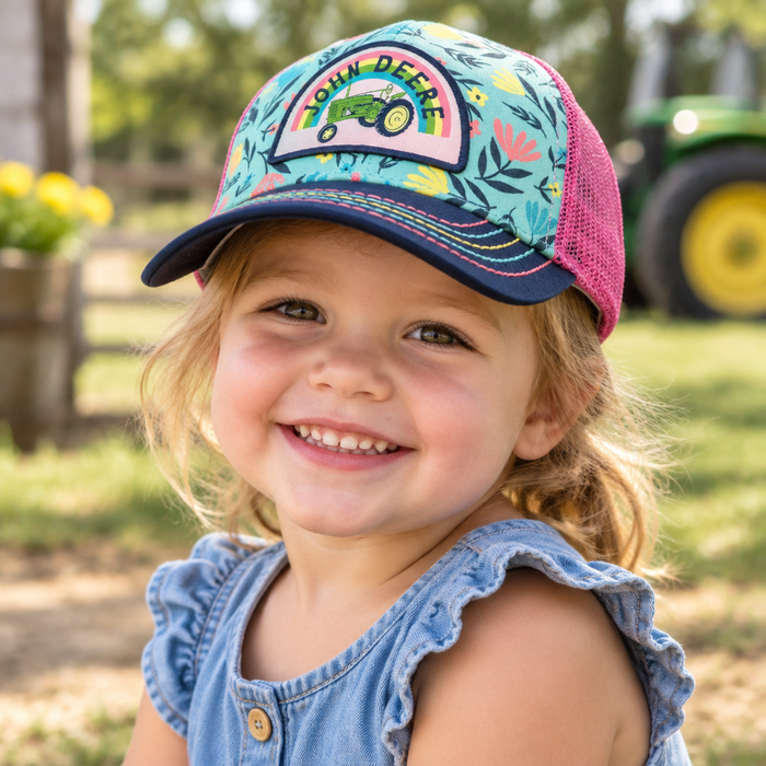 A young girl smiling outdoors while wearing a colorful John Deere trucker hat with a floral pattern and a denim top.
