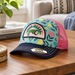 A children's John Deere Toddler Girls Rainbow Tractor Cap resting on a wooden table in a cozy living room setting. The cap features a light blue front panel with a colorful floral pattern, a pink mesh back, a navy blue brim, and a prominent 'John Deere' patch on the front.