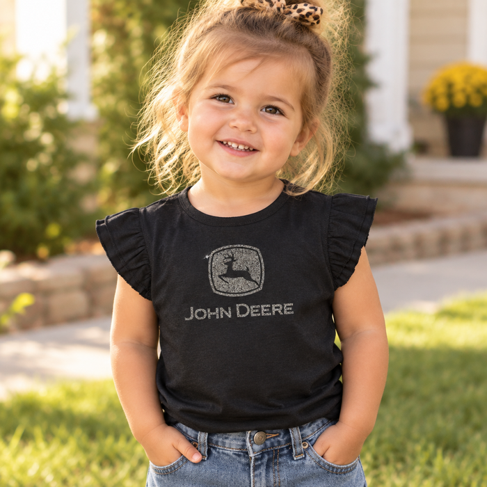 A young girl smiling outdoors while wearing a black short-sleeve t-shirt with ruffled sleeves and a distressed John Deere logo on the front, paired with blue jeans.