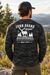 A man wearing a black camouflage ohn Deere Men's Camo Deer & Nature Longsleeve Tee, standing outdoors in a mountain landscape.
