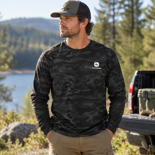 A man wearing a dark gray and black camouflage long-sleeve shirt with a white John Deere logo on the chest, standing outdoors near a lake and trees with a truck in the background.