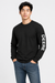 A man wearing a black long-sleeve crewneck shirt with 'DEERE' printed vertically in white on the right sleeve, paired with blue jeans, against a light gray studio background.
