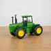 A green John Deere toy tractor with yellow wheels, displayed on a light wooden tabletop against a plain light gray background.