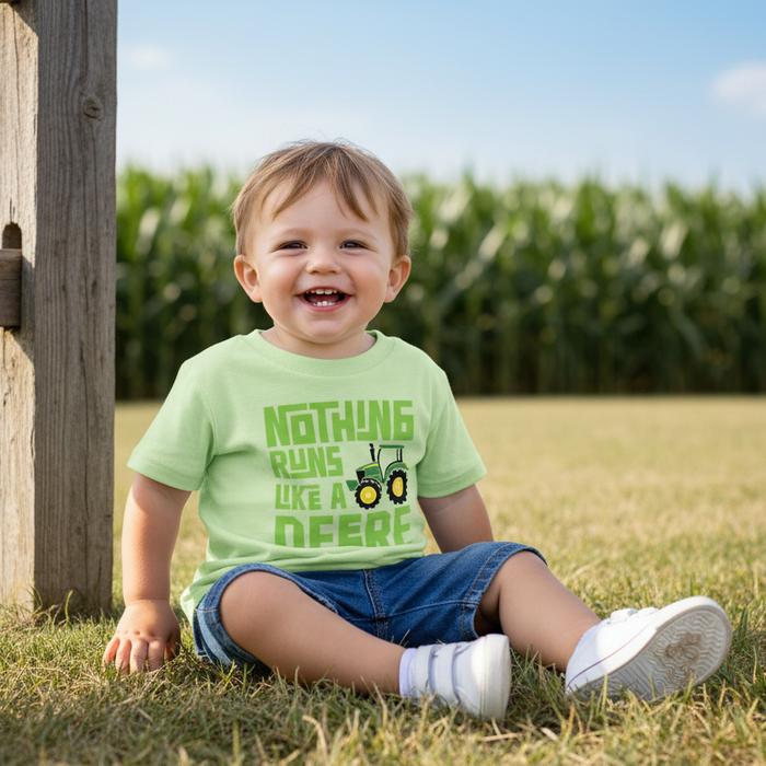 A smiling toddler sitting on grass outdoors wearing a light green graphic T-shirt that reads 'NOTHING RUNS LIKE A DEERE' with a tractor illustration, blue denim shorts, white socks, and white sneakers. A wooden post and blurred cornfield are visible in the background.