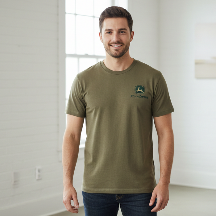 A man wearing an olive green crewneck t-shirt with a small John Deere logo on the left chest, standing in a bright, modern room with white walls.