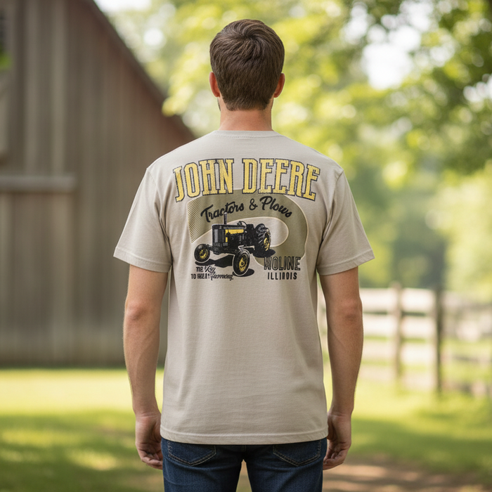 A man viewed from behind wearing a beige t-shirt with a large John Deere graphic on the back, set against a blurred outdoor farm background.